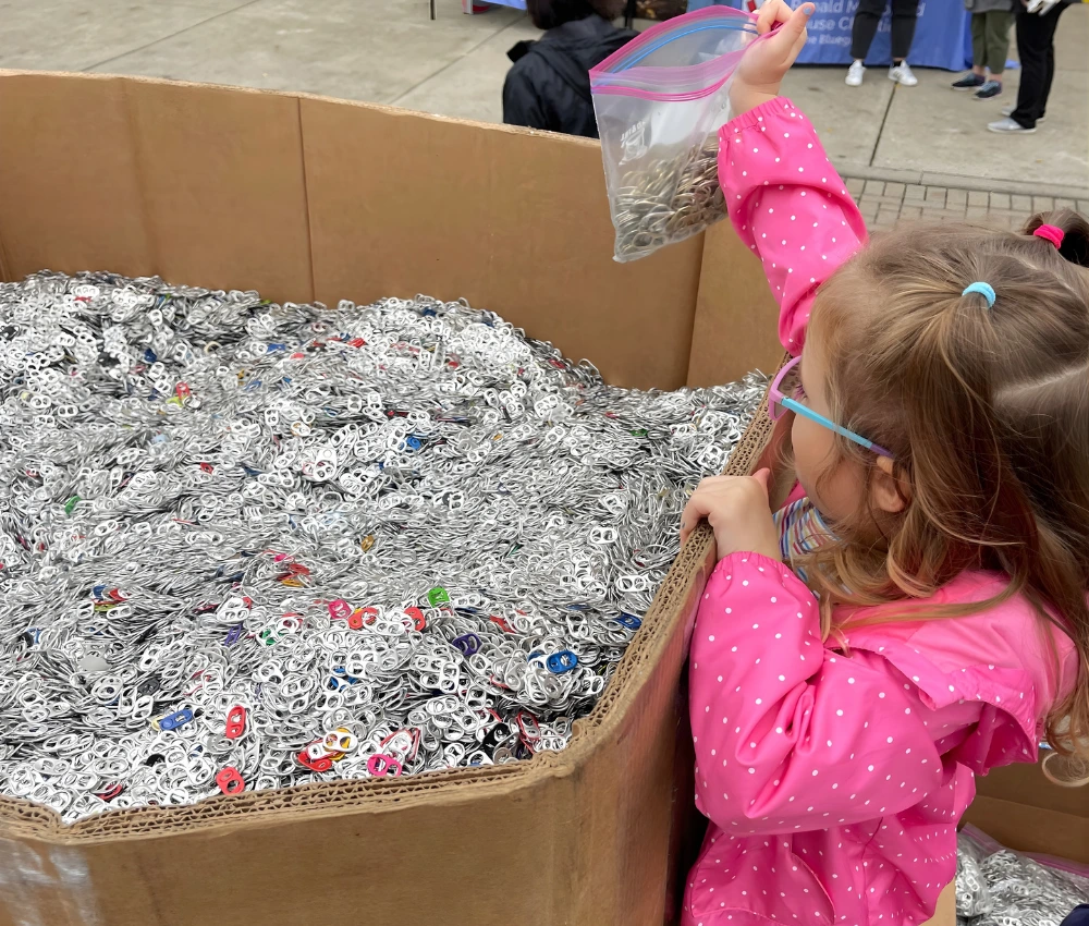 young girl with a bag of soda can pop tabs adding them to a large box of donated pop tabs