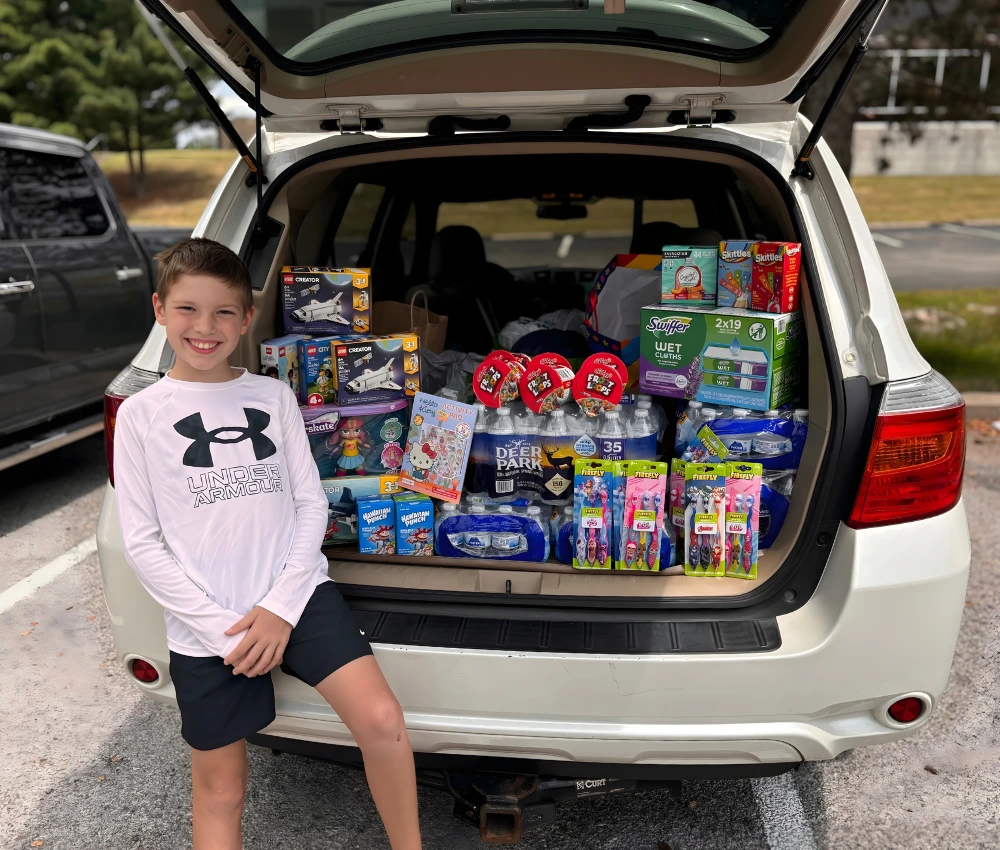 teenage boy sitting against open SUV trunk loaded with goods to donate